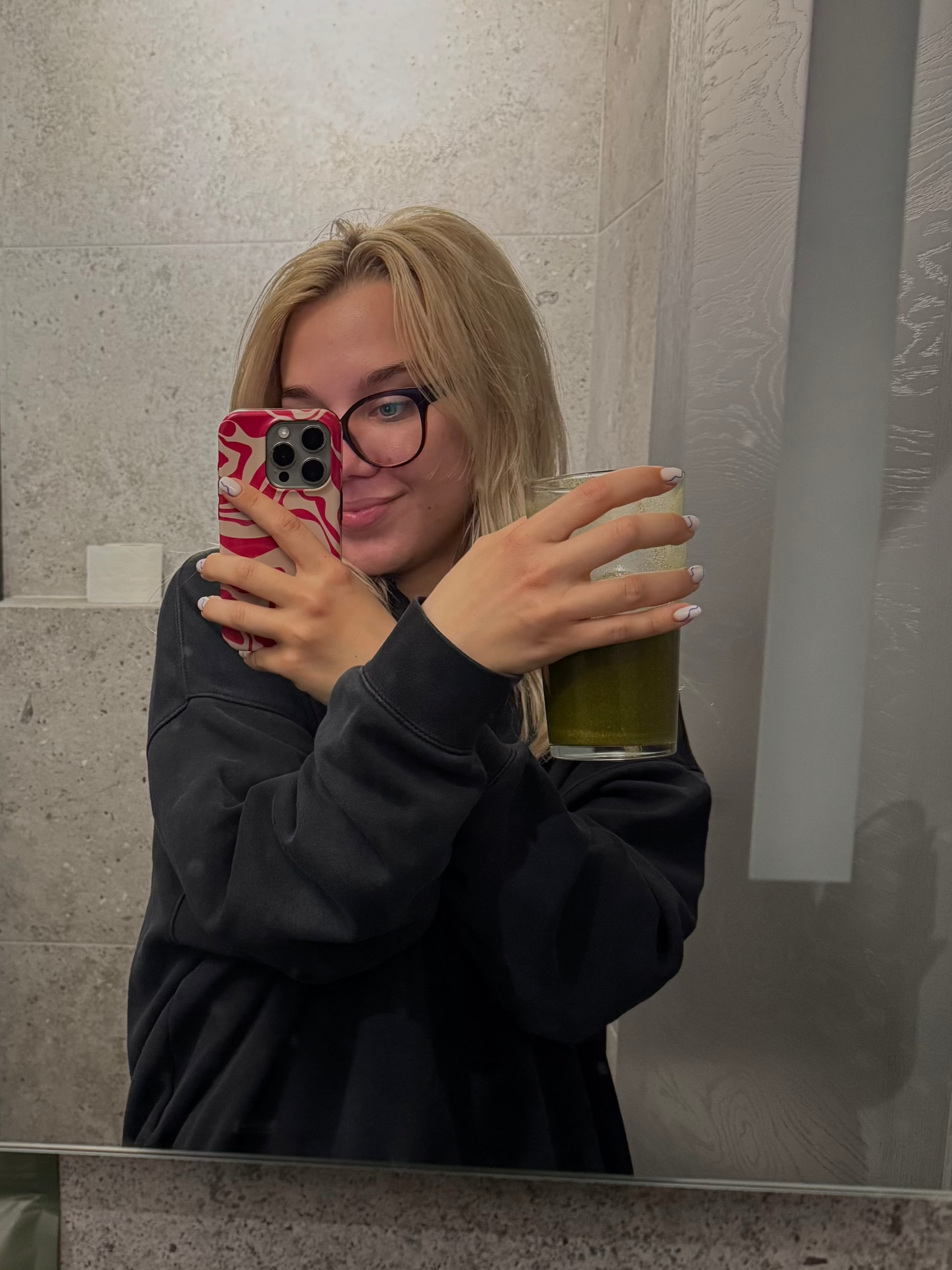 Mergina šviesiais plaukais, laiko stikline su Kapha Blend mišiniu ir fotogorafuoja save žiūrint į veidrodį